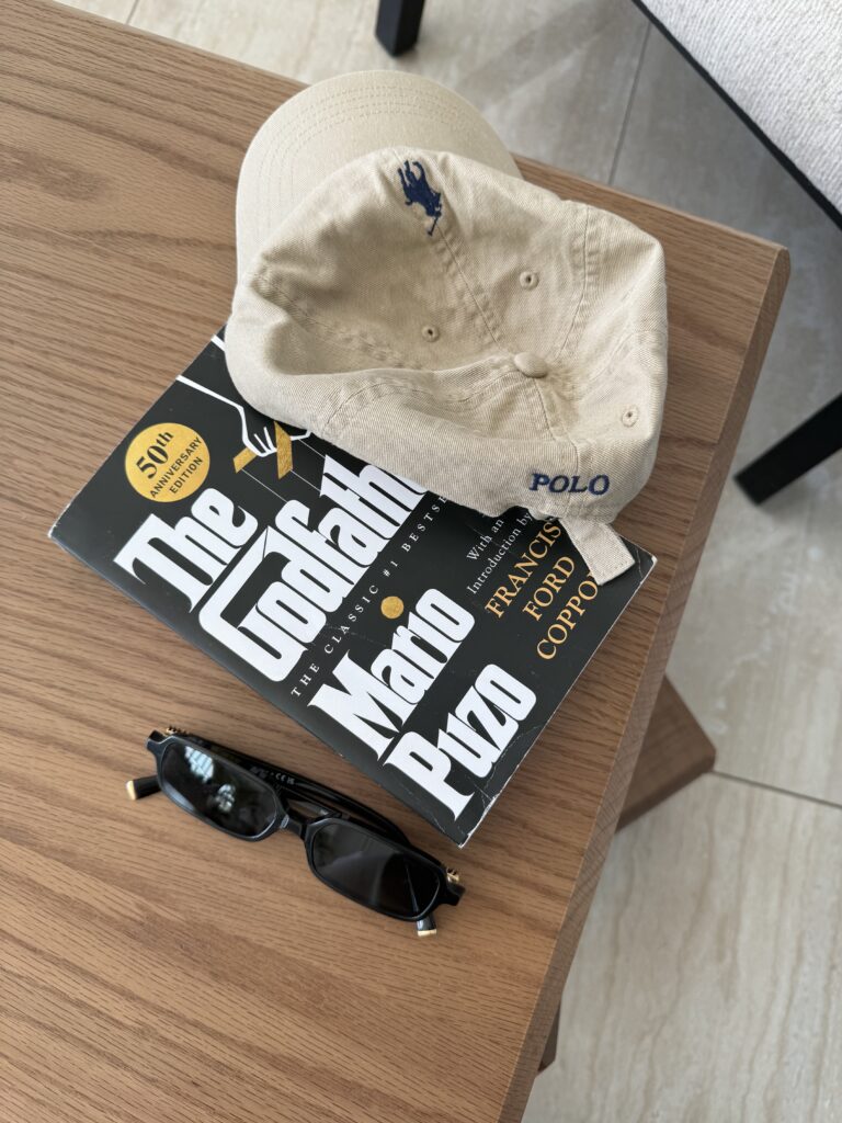 a picture of The Godfather by Mario Puzo book on a wood table with a polo Ralph Lauren hat and sunglasses near it 